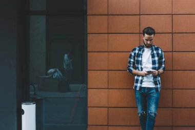 Ciddi bir adam gündelik giysiler giyip akıllı telefon ekranına bakıyor ve bina duvarına yaslanırken mesaj atıyor.