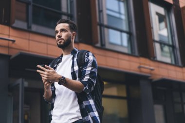 Modern ofis binasının yakınındaki akıllı telefon kullanan, sırt çantalı, rahat giyimli, düşük açılı bir adam.