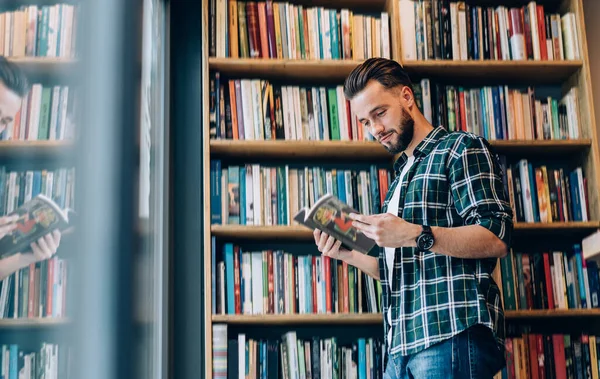 穿着格子衬衫 戴着腕表 留着小胡子的男性站在书架上取书 同时在现代图书馆里呆着 — 图库照片