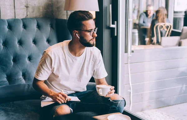 身穿休闲装和眼镜的多愁善感的男性 一边坐在咖啡厅的沙发上 一边看着窗外 一边用无线耳机写诗 一边喝咖啡 — 图库照片
