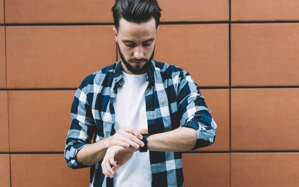 Focused bearded young man in style standing near building wall and looking down at wristwatch checking time outside in daytime