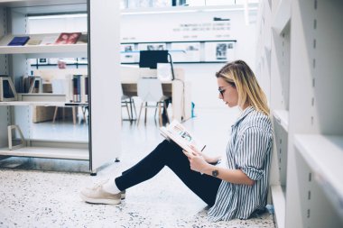 Gözlüklü konsantre hipster öğrenci kütüphanede yerde oturan ve eğitim literatürünü kontrol eden zeki bir kadının yan görüşü olan sınavlara hazırlanırken bilgi okuyor.
