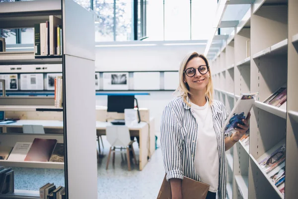 Half length portrait of cheerful youthful woman in optical eyeglasses for vision correction smiling at camera while selecting literature book from bookcase in public library, education concept