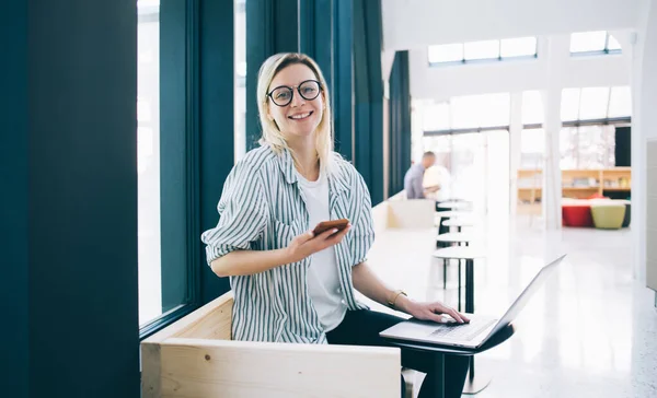 Portrait of cheerful female freelancer in stylish eyewear smiling at camera during mobility networking in coworking space with good internet connection, happy Caucasian blogger enjoying browsing time