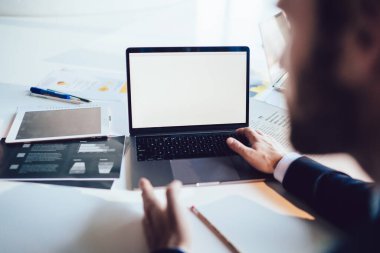 Kağıt çizelgeleri ve kağıt kırtasiyesiyle dolu masada bulanık bir erkek işçi. Ofiste beyaz ekranla laptopta çalışıyor.