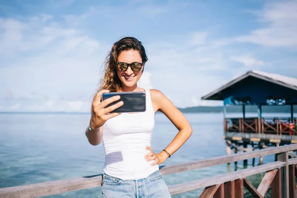 度假期间 站在码头上手挽手 手持手机自拍的戴太阳镜的成年女性微笑 — 图库照片