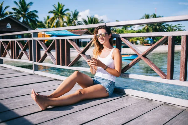 迷人的成年女性穿着太阳镜 白衬衫 牛仔短裤坐在码头上 在暑假期间使用手机 — 图库照片