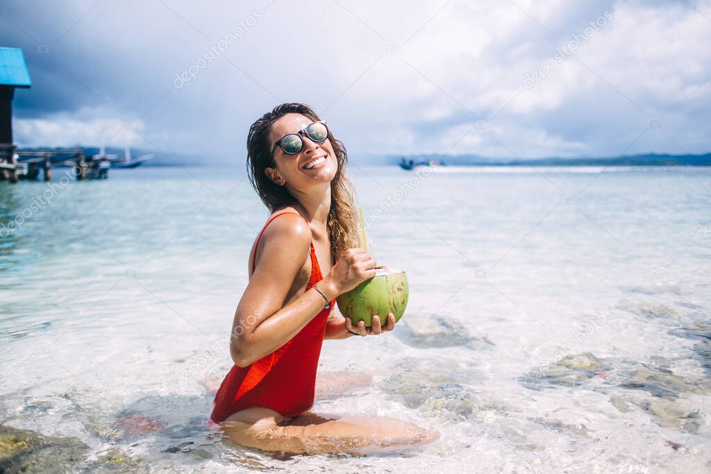 Portrait of funny Caucasian girl in trendy sunglasses refreshing in summer sea holding coconut fruit and smiling at camera, cheerful woman with natural beverage posing at seashore of tropical Cayman