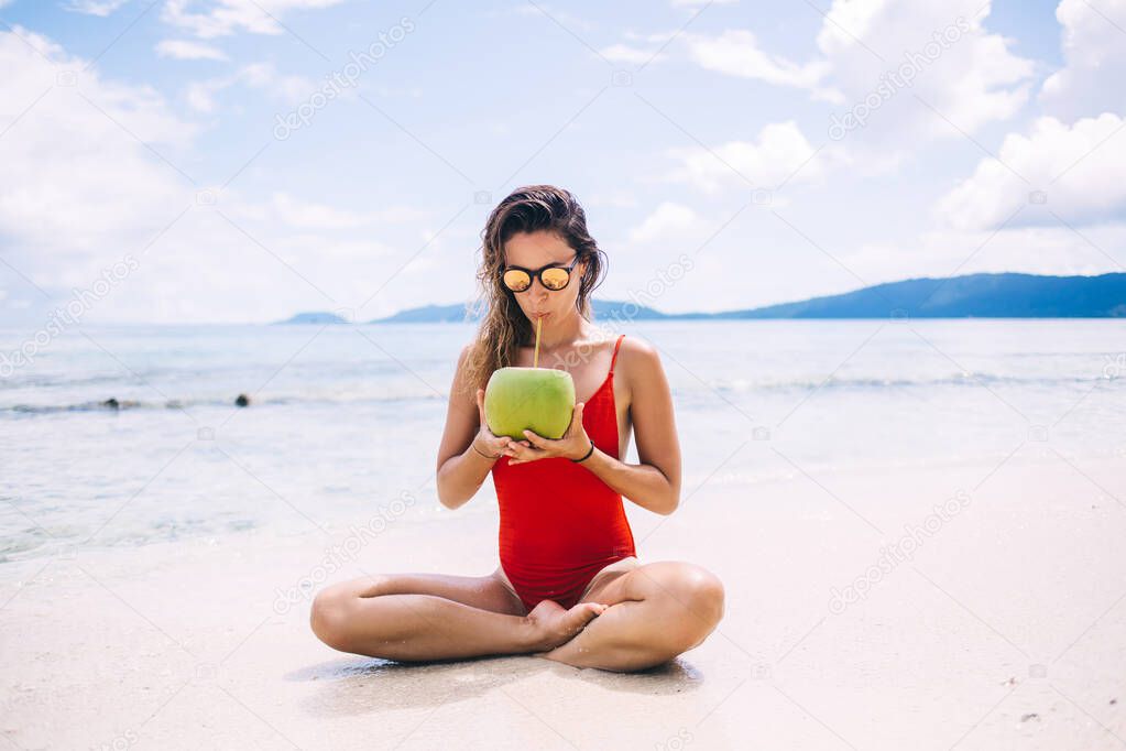 Caucasian woman in red swimsuit drinking coconut beverage during sunbathing on tropical Hawaii beach, young female in sunglasses enjoying healthy vegan food during summer vacations on Seychelles
