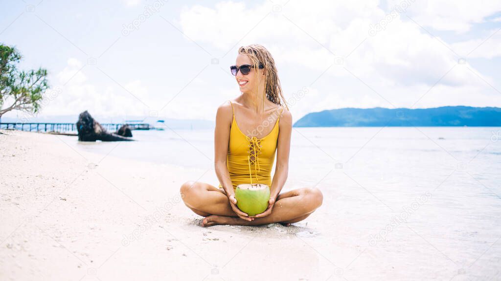 Cheerful female in sunglasses resting at tropical sand beach with coconut fruit satisfied with sunbathing leisure, funny Caucasian woman in sunglasses enjoying vacations on Raja Ampat island