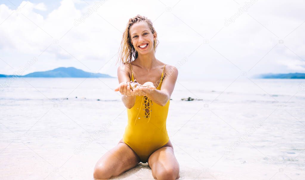 Portrait of cheerful woman in yellow swimsuit posing at sand beach recreating during summer vacations on Seychelles, happy Caucasian female holding shell mollusk enjoying collecting hobby