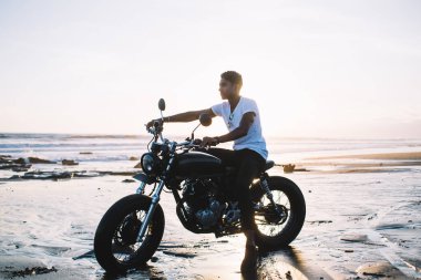 Beyaz tişörtlü ve siyah pantolonlu İspanyol bir adamın kumlu okyanus kıyısında, gün doğumunda bulanık arka planda otururken ve gidonu tutarken önüne baktığı yan görüntü.