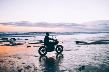 Tanımlanamayan, düşünceli, riskli bir adamın, bulanık arka planda romantik günbatımında deniz kenarında hızlı, çağdaş bir motosiklet sürüşünün yan görüntüsü.