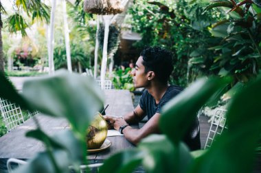 Yan bakış açısıyla, günlük giysiler içindeki konsantre etnik erkek sokak kafesindeki ahşap masada dinlenirken deftere not alıyor ve serinletici hindistan cevizi içeceklerinin tadını çıkarıyor.