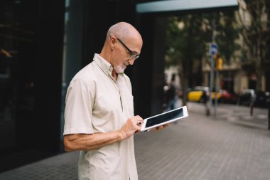 Şehir caddesinde kaldırımda dikilip tablet kullanan günlük giysiler giymiş sakallı bir erkek.