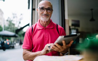 Modern dokunmatik bloknotu elinde olan, boş zamanlarında kameraya gülümseyen, günlük pembe tişört giymiş, dijital tablet üzerinde 4 g internet kullanan olgun, beyaz bir adamın yarım boy portresi.