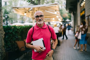 Seyahat haftasonu kameralarına bakarak göz düzeltmesi yaptığı için beyaz gözlüklü yaşlı insan, dijital cihaz tutan ve şehir caddesinde poz veren yaşlı blog yazarı