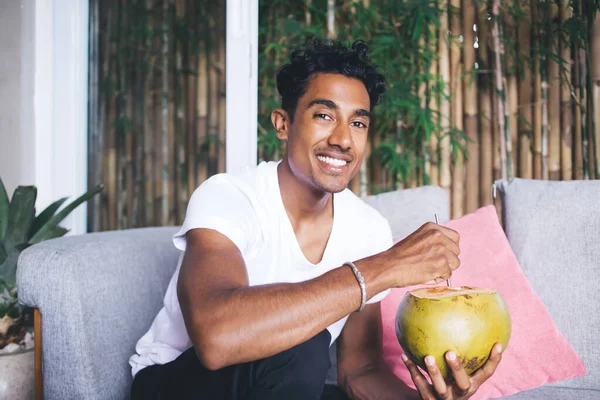 穿着休闲装的西班牙裔男子坐在热带自助餐厅的沙发上 面带微笑 喝着新鲜椰子汁和稻草 — 图库照片