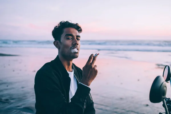 日落黄昏时分 身穿深色外套的忧郁的西班牙裔男子拿着香烟 在海滨波涛汹涌的水面上若有所思地喷出浓烟 — 图库照片