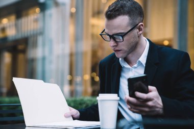 Gözlüklü ve takım elbiseli genç adam kaldırımdaki kafede oturuyor ve arka planda akıllı telefon tutarken iş gazetelerini okuyor.