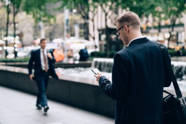 Gözlüklü, siyah çantalı, sokakta duran ve arka planda gülümseyen akıllı telefon kullanan müdürün resmi görünümü