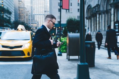 Siyah iş ceketli, gözlüklü, omuz çantalı ve telefon kullanırken caddeden karşıya geçen çekici adamın yan görüntüsü.