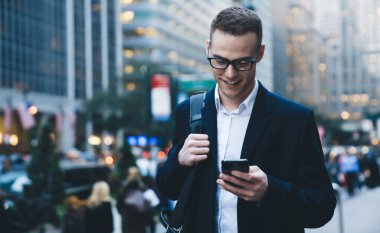Resmi giyinen ve moda gözlüklü genç girişimci şehir merkezinde sokakta yürürken ve çanta taşırken gülümsüyor ve akıllı telefonlara bakıyor.