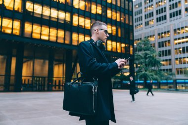 Sosyal medyada çantalı ve kulaklıklı bir adamın sokakta dikilip gözlerini kaçırırken akıllı telefon kullanarak sohbet edişinin yan görüntüsü.