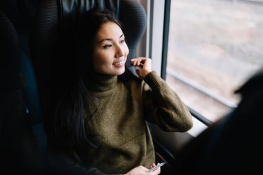 Sıradan giyinmiş gülümseyen Asyalı kadın gezgin tren yolculuğunda rahat bir vagonda oturmuş pencereye bakıyor, modern tren yolculuğundan memnun kalmış güzel bir kadın turist.