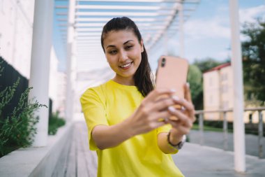 Sarı tişörtlü genç Asyalı kadın güzel bir yaz gününde sokakta durup cep telefonundan selfie çekiyor.