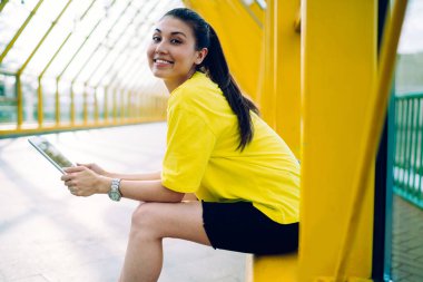 Normal kıyafet ve kol saati giymiş, dişlek gülümsemeli, sarı metal korkuluklara oturmuş kameraya ve tablete bakarken pozitif etnik kadın görüntüsü.