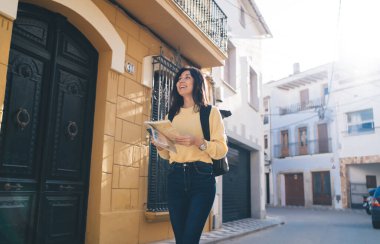 Sıradan giysiler içinde, sırt çantalı, şarjörlü ve kaldırımda yürürken yüzünü çeviren neşeli, etnik bir kadın.