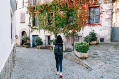 Köyde antik mimari ve yeşillik içinde yürüyen genç turistlerin arka plandaki manzarası. Seyahat sırt çantalı genç hippi kız tatillerde gezintiye çıkıyor.