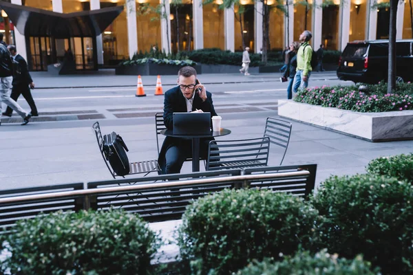 Focused male entrepreneur in formal attire and eyeglasses talking on smartphone and working on tablet while sitting on downtown street with coffee