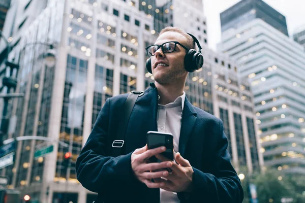 From below young male entrepreneur in formal attire and glasses standing in downtown while listeinging to music from smartphone through headphones