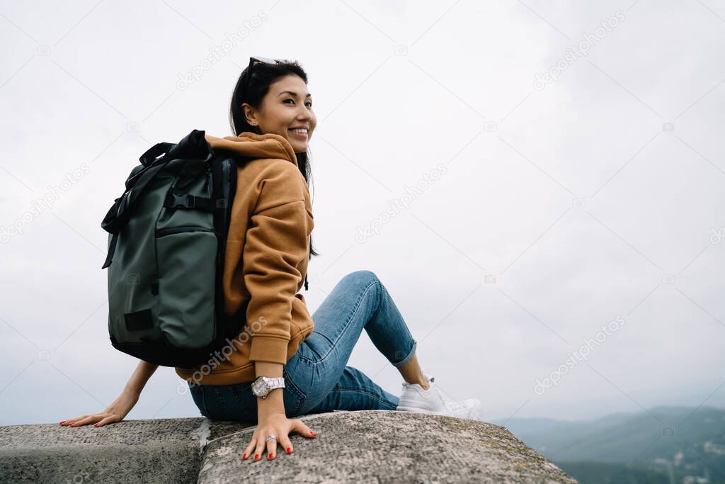 Smiling asian female traveler sitting on destination during sightseeing tour enjoying vacation wanderlust journey, beautiful woman tourist with backpack recreating on exploring route on historic place
