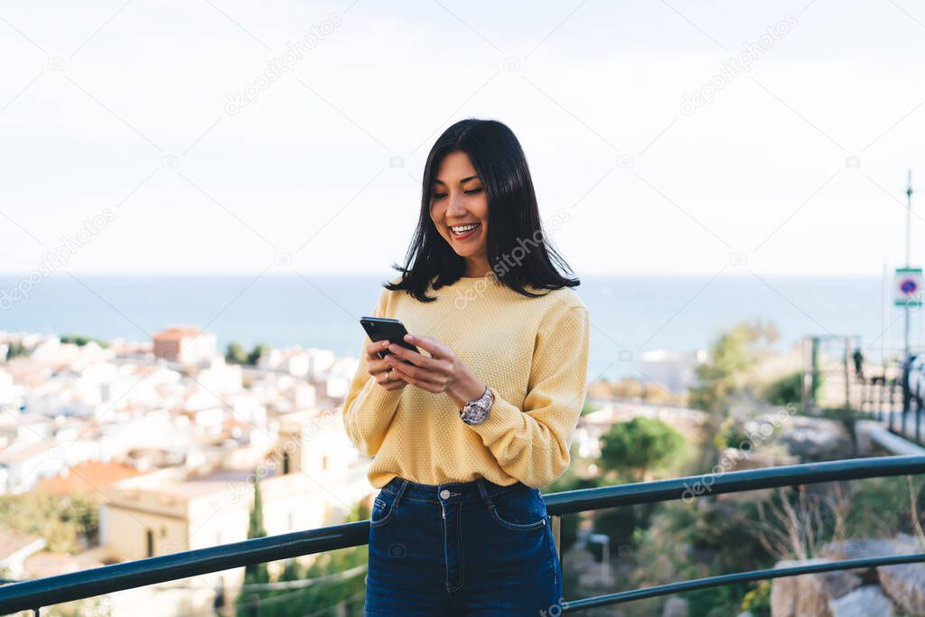 Cheerful Asian hipster girl enjoying smartphone networking during travel vacations for exploring city using good internet connection at overlooking area, joyful female vlogger messaging with followers