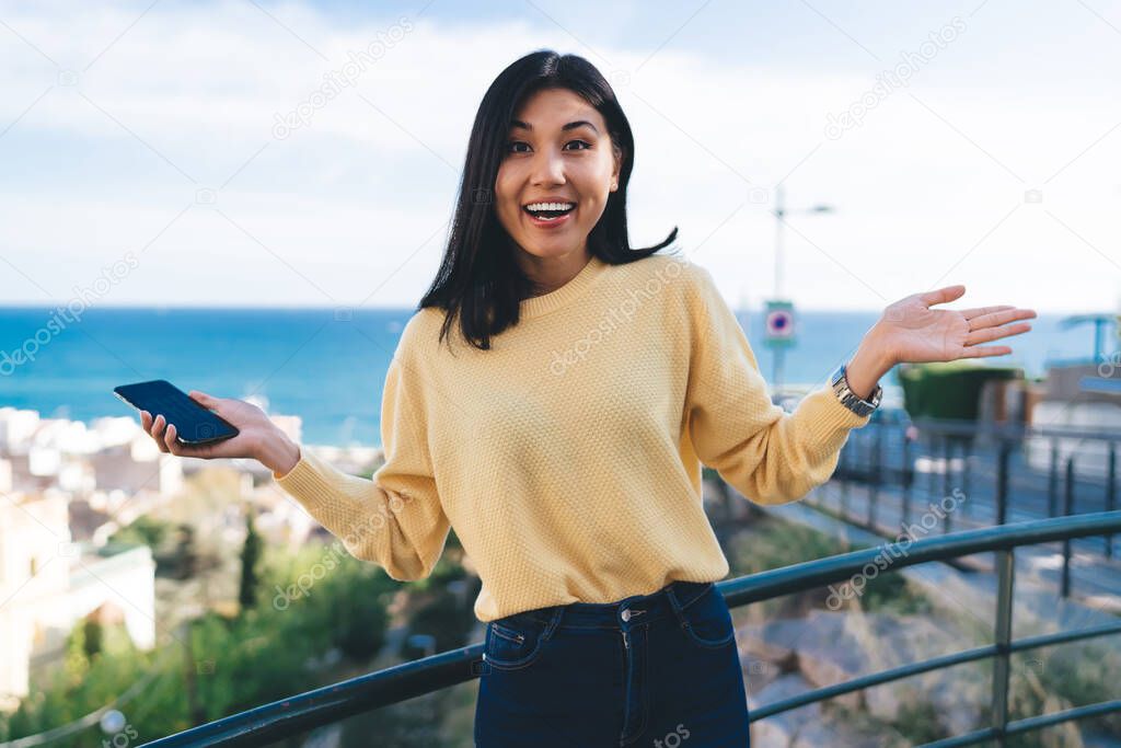 Half length portrait of joyful hipster girl with cellular technology in hand smiling at camera during travel sightseeing at overlooking area, cheerful female traveller shrugging in touristic town