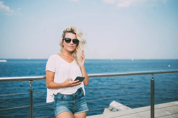海辺の桟橋で夏の遊歩道のための時間の間にカメラを見て流行のサングラスで千年の女性の半分の長さの肖像画 白人ヒップスター女の子は現代のスマートフォン技術を保持 — ストック写真