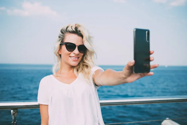 快乐的女性游客享受着散步的快乐时光 在蓝色大海的背景下点击了自拍的照片 戴着太阳镜的无忧无虑的嬉皮士女孩使用现代手机拍摄博客 — 图库照片