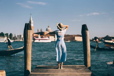 Venedik, İtalya 'da rüzgarlı bir günde ahşap iskelede dinlenirken mavi elbiseli ve şapkalı bayanın arka görüntüsü