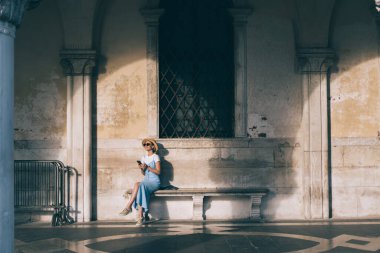 Venedik 'teki kemerli galeride otururken şapkalı neşeli genç bir kadın dinlenirken elinde akıllı bir telefon tutuyor.