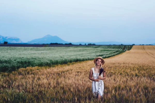 Half length portrait of happy woman with flowers bouquet posing in barley field and smiling at camera during solo journey to rural countryside, pretty female with posy discovering nature environment