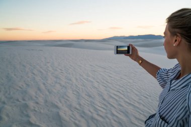 Akıllı telefon kullanan, kum tepeciklerinin fotoğrafını çeken ve White Sands Ulusal Parkı 'ndaki çölü keşfederken gün batımının tadını çıkaran bir doğa yürüyüşçüsü.