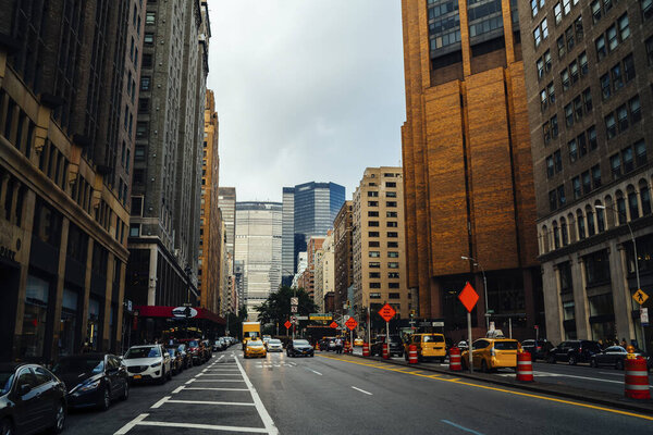 Cars driving on asphalt road near modern high buildings on busy street of New York City in America against gray sky
