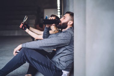 Spor giyim sektöründe ciddi sakallı bir adam spor salonunda antrenman yaptıktan sonra şişeden gözleri kapalı su içiyor.