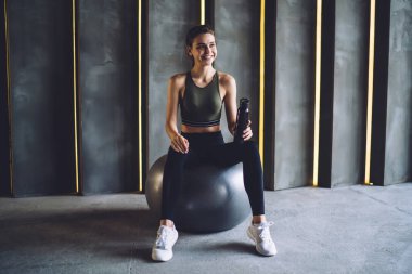Spor kıyafetli, sıska, gülümseyen bir kadın vücudu bir şişe suyla top üzerinde oturuyor ve spor salonunda gözlerini kaçırıyor.