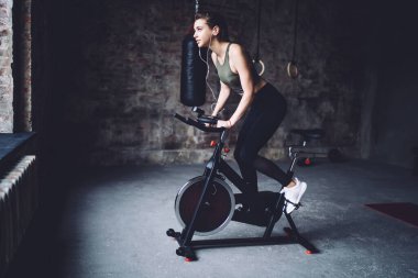 Kolunda cihaz ve kulaklığı olan, spor salonunda sabit egzersiz bisikleti üzerinde kardiyo antrenmanı yapan genç bir kadının yan görüntüsü.