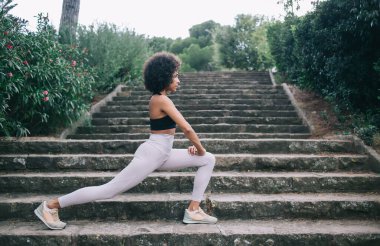 Spor giyimli, spor ayakkabılı ve spor ayakkabılı, yeşil parktaki merdivenlerde Crescent Lunge pozu veren esnek genç bir kadının yan görüntüsü.