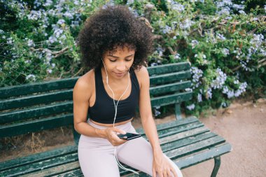 Aktif çalışan genç siyah bir kadın cep telefonu takıyor ve bahçede bankta otururken kulaklıkla müzik dinliyor.